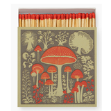 A matchbox filled with red-tipped wooden matches, featuring a vintage-style illustration of red mushrooms and botanical motifs on a muted gray-green background.