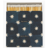 A box of matches with a dark navy blue cover decorated with a repeating cream star pattern and gold text reading 'luxury matches'. The wooden matches with white tips are visible at the top.