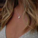 A close-up lifestyle shot of a woman wearing a delicate silver chain necklace with a small daisy pendant. The pendant features a white center and yellow petals, worn against her collarbone over a light gray floral top.