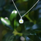 A delicate silver chain necklace with a small white daisy pendant featuring a yellow center, photographed against a blurred background of green leaves.